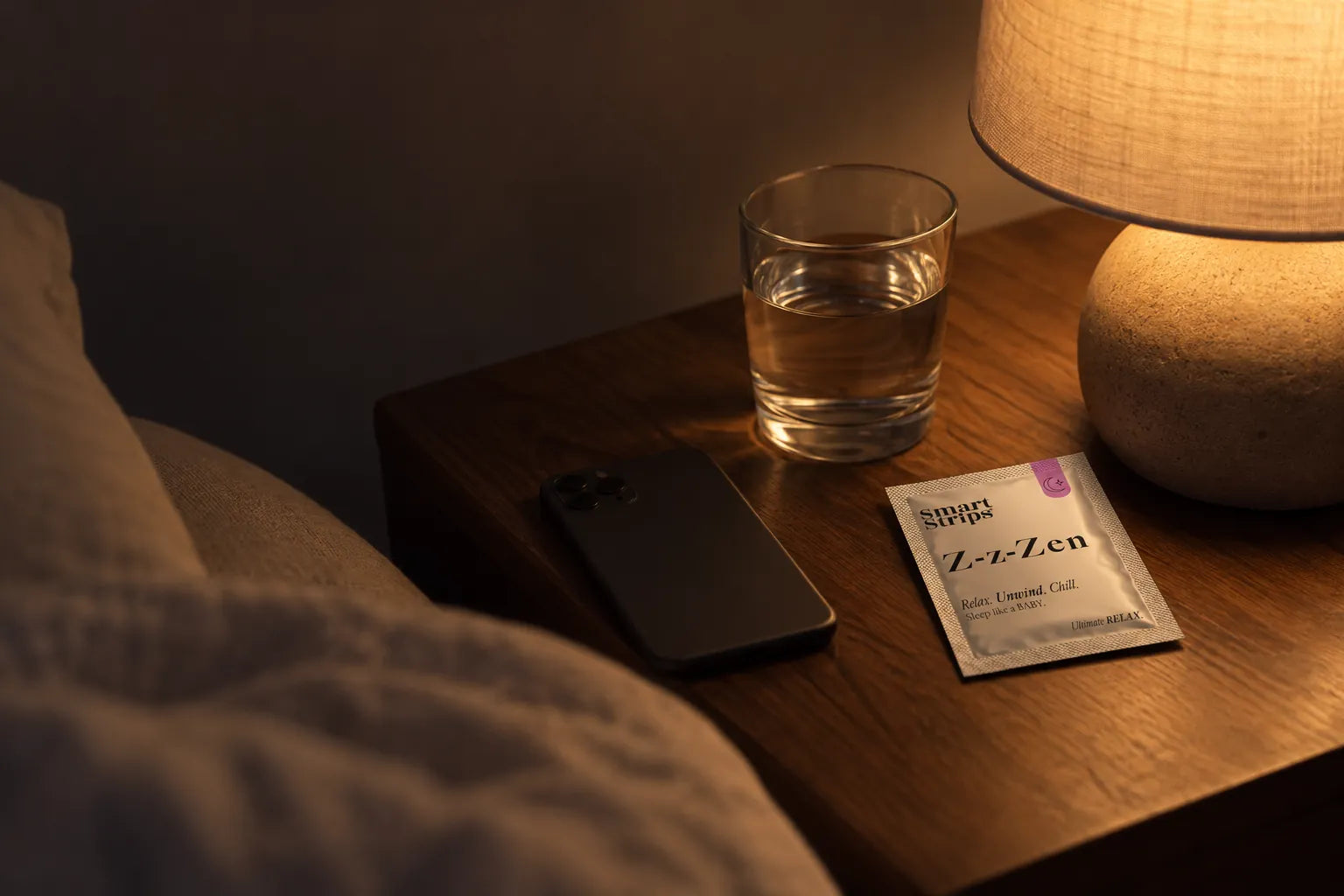 Bedside table in warm lamp light with a glass of water, a phone face down and a Smart Strips Z-z-Zen sachet